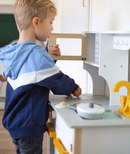 Dziecko bawiące się białą i złotą drewnianą kuchnią Tryco z otwieranymi drzwiczkami i akcesoriami kuchennymi