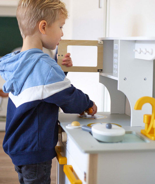 Dziecko bawiące się białą i złotą drewnianą kuchnią Tryco z otwieranymi drzwiczkami i akcesoriami kuchennymi